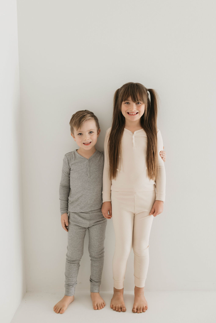 Two children stand against a white wall, smiling at the camera. The child on the left wears the "Bamboo Two Piece Pajamas" in Ribbed Grey from forever french baby, while the child on the right wears a light pink top and pants. The child on the right has long hair styled in pigtails.