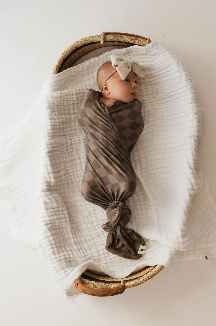 An infant wrapped in the forever french baby's Bamboo Swaddle | Faded Brown Checkerboard sleeps peacefully in a wicker basket lined with a white, textured blanket. The baby wears a white bow headband.