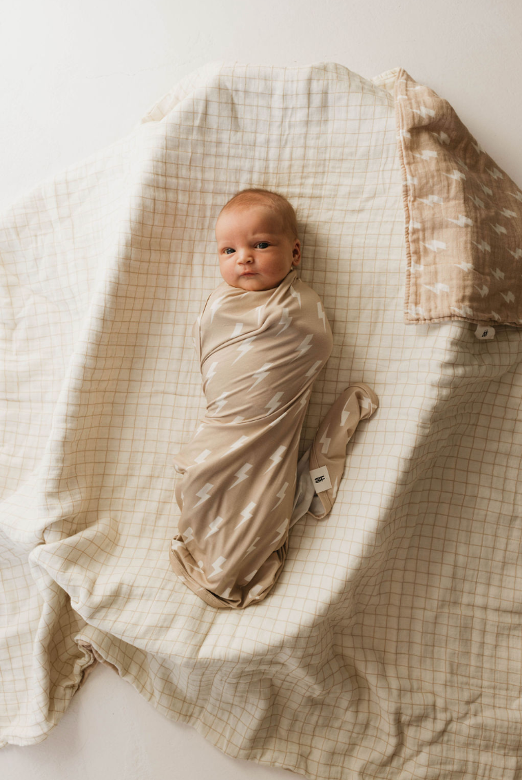 A baby is swaddled in the forever french baby's Bamboo Swaddle featuring a tan and cream lightning bolt pattern, lying on a beige and white checkered blanket. There is another light brown patterned blanket partially visible beside the baby. The baby has a calm expression and is looking up.