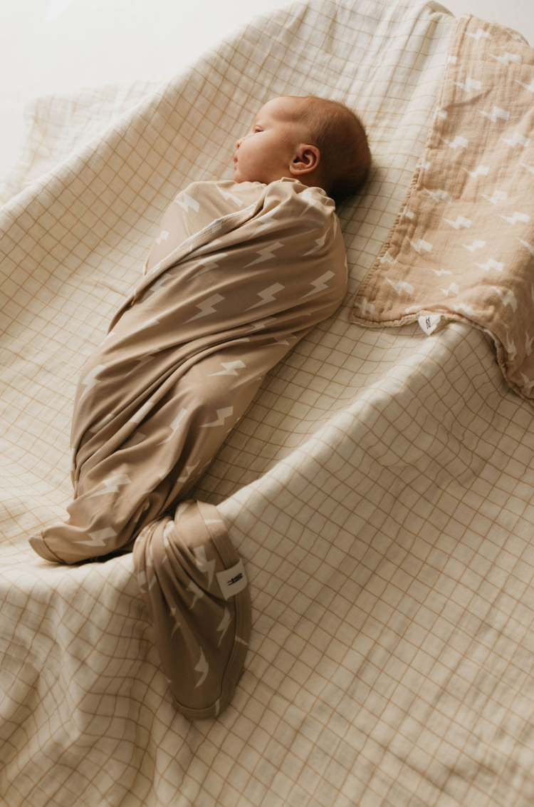 A baby sleeps peacefully, swaddled in a hypo-allergenic Bamboo Swaddle in Tan & Cream Lightning Bolt pattern from forever french baby, lying on a cream-checkered cloth. Another Bamboo Swaddle with the same pattern is folded nearby. The baby is covered from neck to feet, with only the head visible, suggesting a cozy and secure environment.
