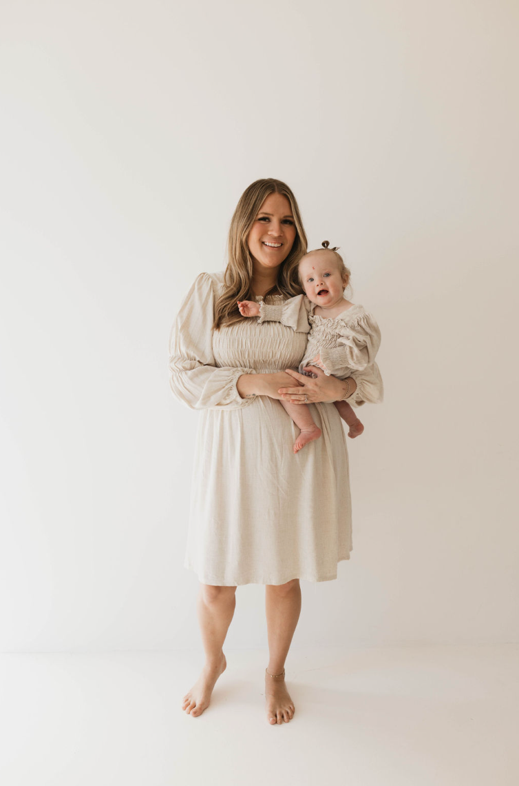 A woman with long brown hair, wearing a luxurious flax-colored linen dress, stands barefoot holding a baby dressed in the "Linen Romper | Flax" from forever french baby. Both are smiling against a plain white background, and the baby, adorned with a small bow in her hair, looks adorable in her outfit available in sizes 0-24 months.