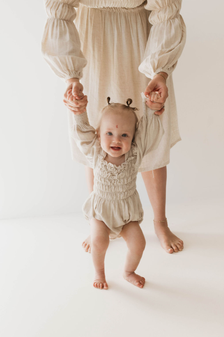 A happy baby, dressed in a chic Linen Romper in Flax from forever french baby, holds onto an adult's hands for support while standing. The adult, also clad in beige and partially visible in the background, helps the baby walk on a pristine white floor. Both are barefooted. This luxurious flax-colored linen romper is available in sizes 0-24 months.