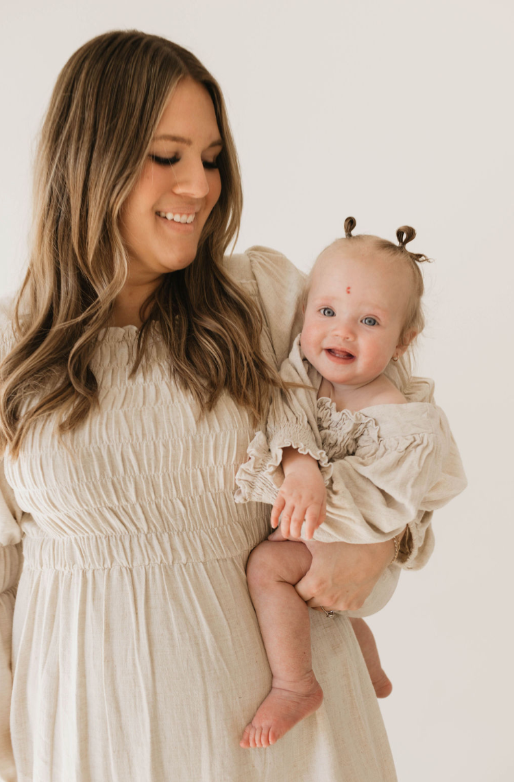 A woman with long, wavy hair wearing a beige dress holds a smiling baby dressed in the luxurious, flax-colored Linen Romper by forever french baby. The baby, with two small ponytails and a mark on the forehead, radiates joy. Their matching outfits create an endearing scene against a plain, light-colored background.