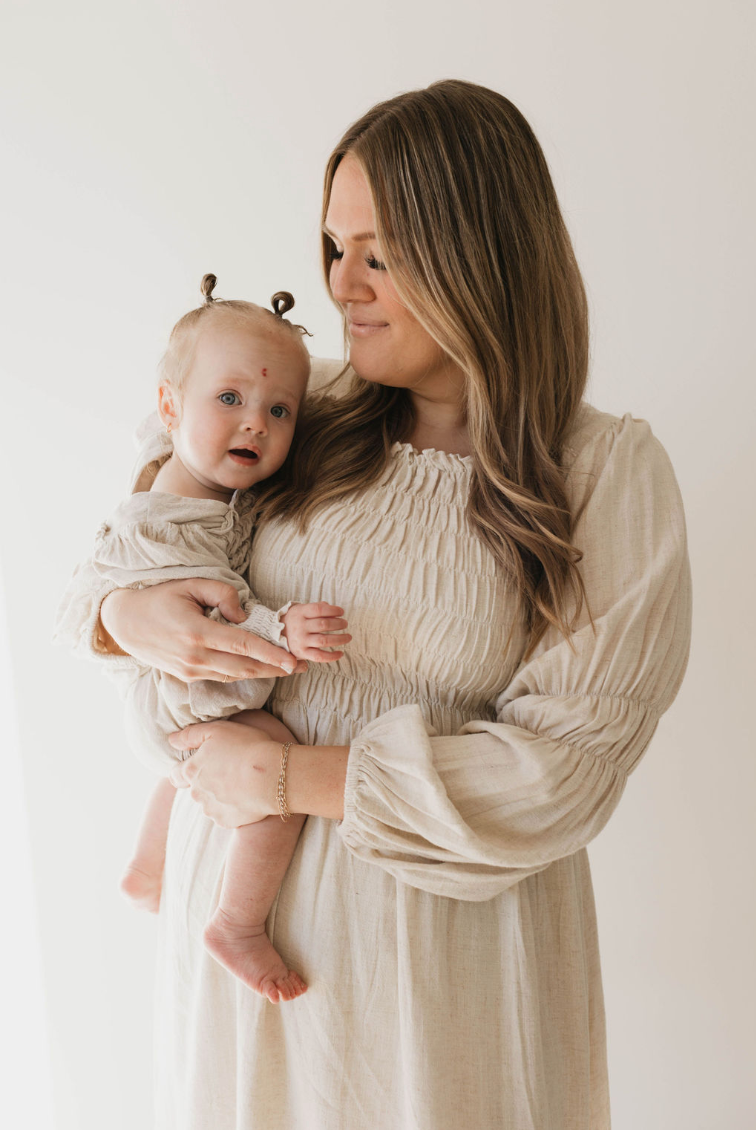 A woman with long blonde hair, dressed in a luxurious flax-colored linen gown, cradles a baby who is wearing the forever french baby's Linen Romper in Flax, available in sizes 0-24 months. They both gaze softly to the side, standing against a plain white background. The woman holds the baby tenderly in her arms.