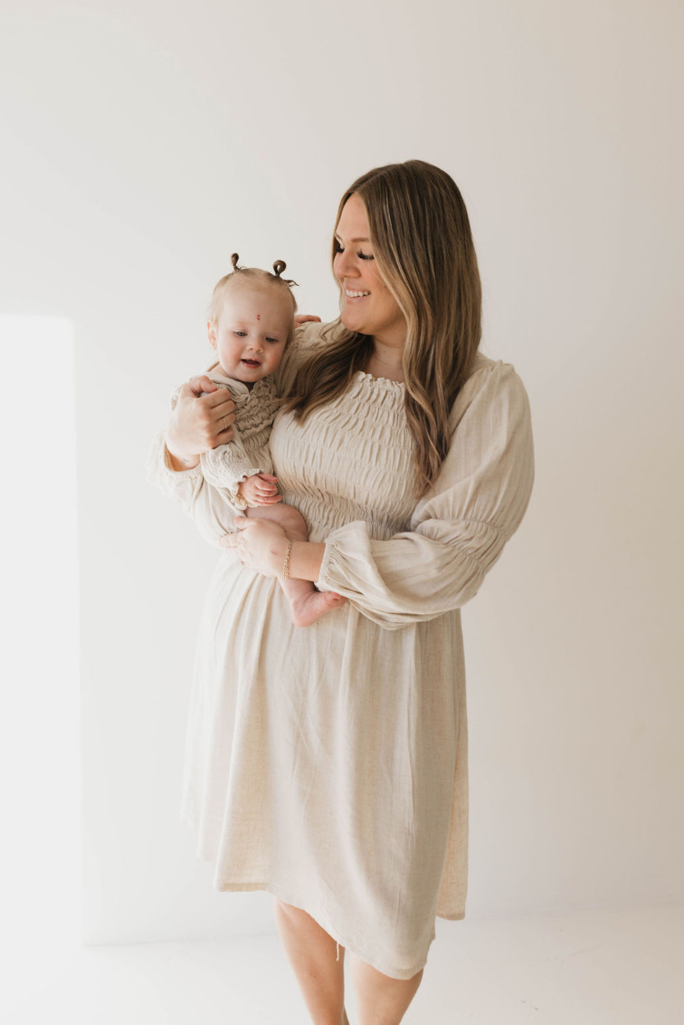 A woman with long hair, wearing a light-colored dress, is holding a baby dressed in a forever french baby Linen Romper in Flax. This luxurious linen outfit, suitable for sizes 0-24 months and paired with the baby's small pigtails, complements the warmth and affection in their smiles as they stand against a simple white background.