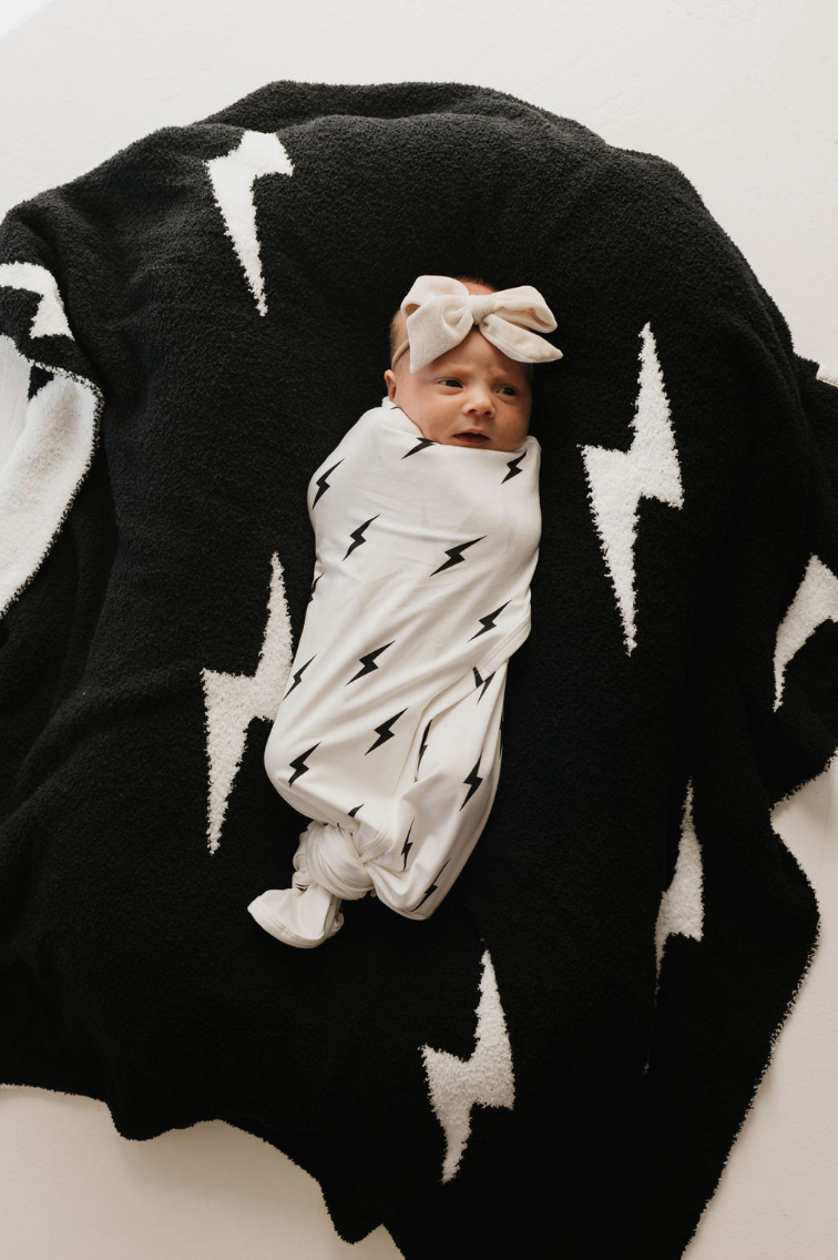 A baby, swaddled in the Bamboo Swaddle | White & Black Lightning Bolt by forever french baby, lies on a black blanket adorned with white lightning bolt designs. The baby is wearing a bow headband and looking up with a calm expression.