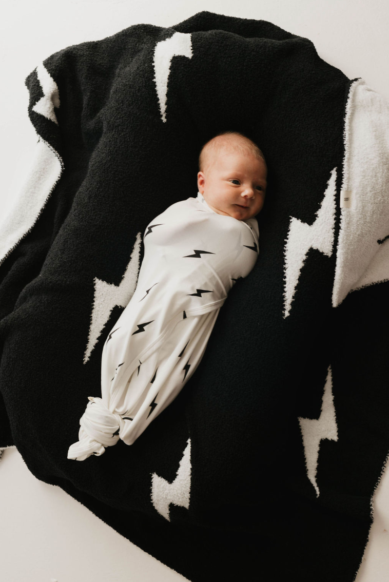A newborn baby is swaddled in a hypo-allergenic Bamboo Swaddle | White & Black Lightning Bolt from forever french baby, lying on top of a black and white blanket with similar designs. The baby is looking slightly to the side.
