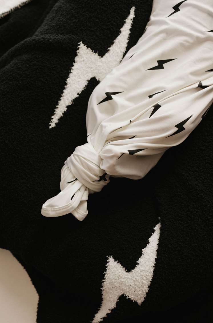 A white fabric adorned with black lightning bolt patterns, similar to the "Bamboo Swaddle | White & Black Lightning Bolt" by forever french baby, is tied in a knot and displayed on a black textured fabric featuring large white lightning bolt designs. The contrasting colors and patterns create a striking visual effect, ideal for hypo-allergenic clothing or breathable baby sleepwear.