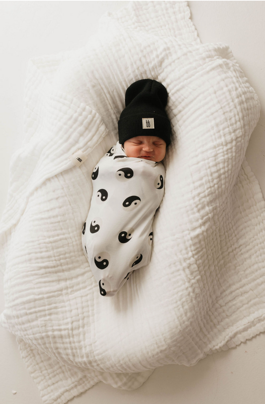 A sleeping baby swaddled in a Bamboo Swaddle | Black & White Yin Yang by forever french baby rests on a crumpled white quilt. The baby is wearing a black knit hat, and the background is plain white.