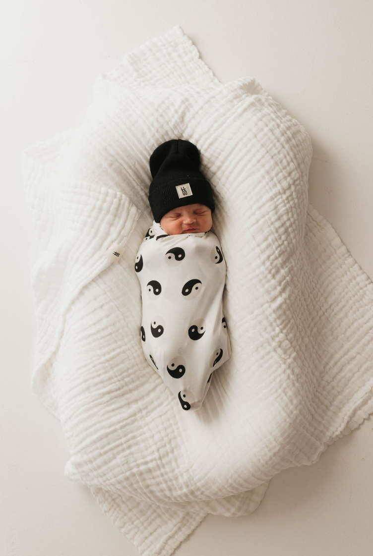 A newborn baby is enveloped in a hypo-allergenic, breathable blanket named Bamboo Swaddle | Black & White Yin Yang by forever french baby, featuring black yin-yang symbols. The baby is also sporting a black beanie hat and is resting on a white, textured, crumpled blanket. The baby's eyes are closed, and they appear to be peacefully sleeping.