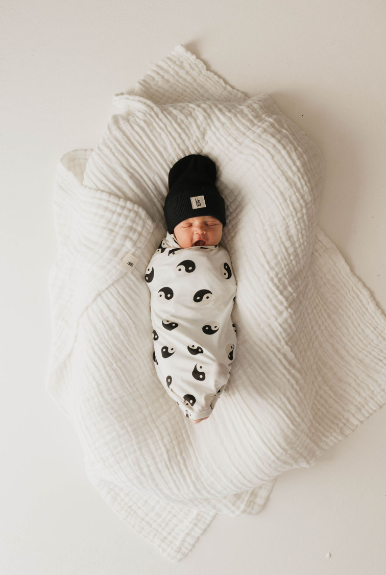A newborn baby is swaddled in a hypo-allergenic forever french baby Bamboo Swaddle | Black & White Yin Yang. The baby lies on a large white quilt, wearing a black knit hat, against the plain white background.