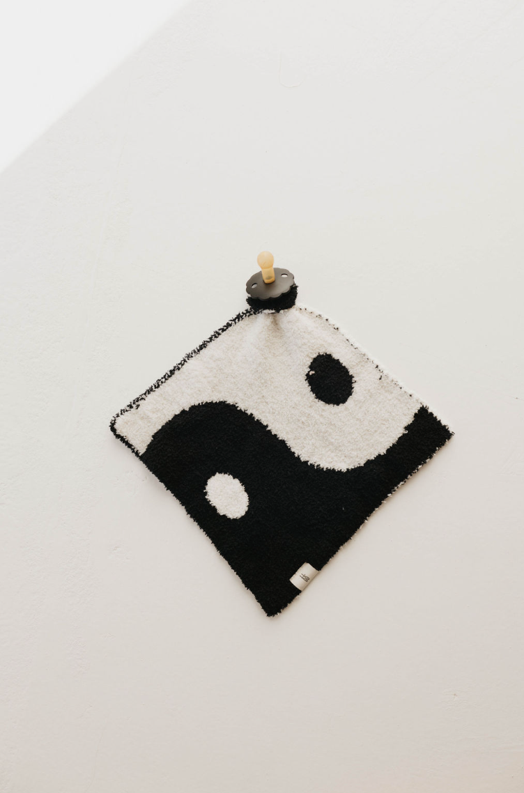 A square Lovey | Yin-Yang ☯︎ towel by forever french baby, featuring a classic black and white yin-yang design, hangs on a white wall, attached by a round wooden peg at the top center. The towel's left side is black with a white dot, and the right side is white with a black dot. It also doubles as a convenient pacifier holder for your little one.