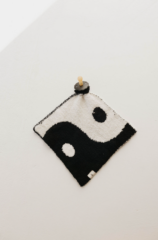A square Lovey | Yin-Yang ☯︎ towel by forever french baby, featuring a classic black and white yin-yang design, hangs on a white wall, attached by a round wooden peg at the top center. The towel's left side is black with a white dot, and the right side is white with a black dot. It also doubles as a convenient pacifier holder for your little one.