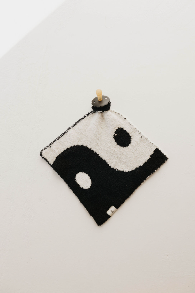 A small Lovey | Yin-Yang ☯︎ wall hanging from forever french baby is displayed against a white wall, featuring a design resembling the Yin Yang Lovey symbol. The hanging is attached to the wall using a circular black peg with a wooden knob at the center.