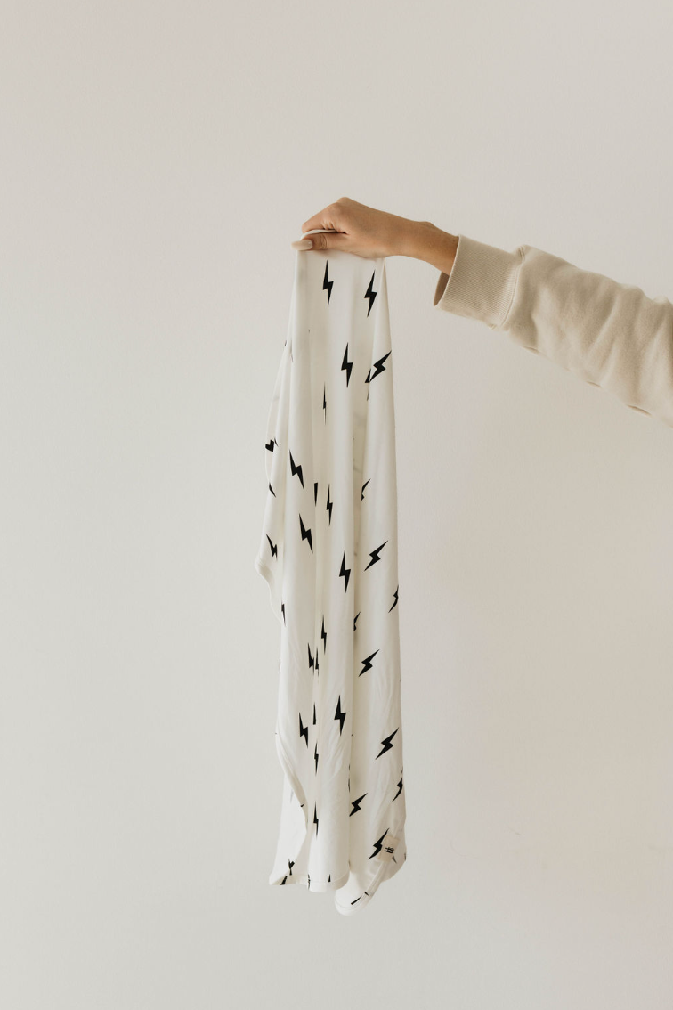 A hand holds the "Bamboo Swaddle | White & Black Lightning Bolt" from forever french baby against a plain beige background. The arm, clad in a long beige sleeve, extends as the breathable baby sleepwear drapes down, perfectly showcasing the full design.