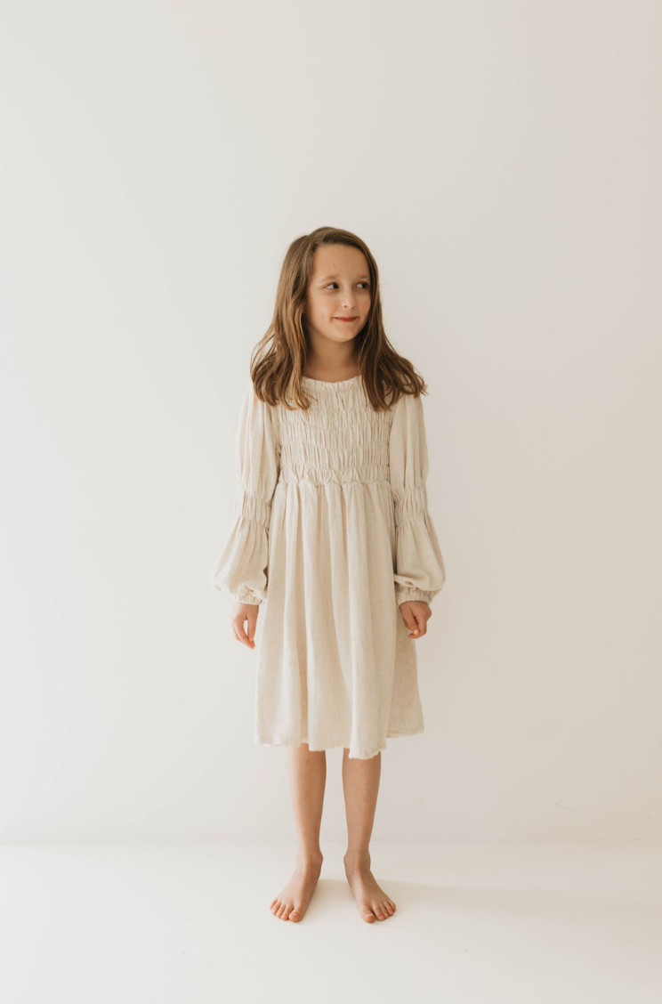 A young girl with long brown hair, dressed in a luxurious "Linen Romper | Flax" by forever french baby, stands barefoot against a plain white background. She is looking slightly off to the side with a calm expression. Available in sizes 0-24 months.