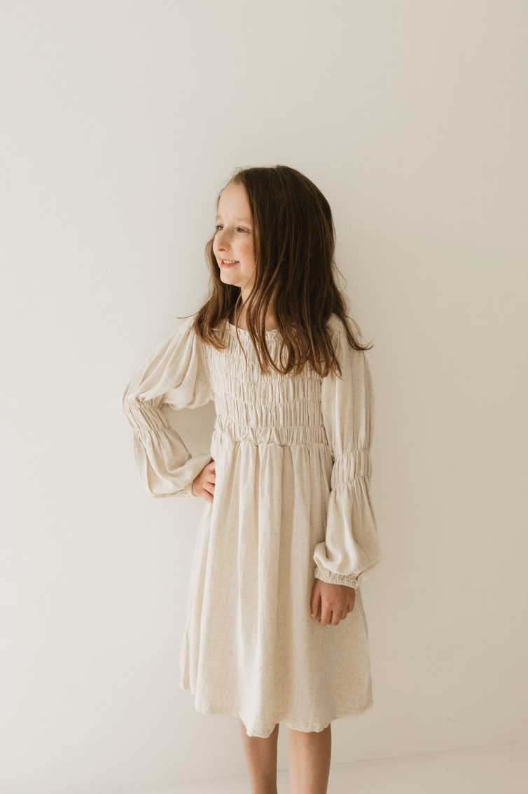A young girl with long brown hair stands against a plain white background, looking to her left and smiling. She is wearing the Women's Linen Dress | Flax from forever french baby, featuring a gathered bodice. Her left hand rests on her hip while her right arm hangs by her side, showcasing the versatile and comfortable design.