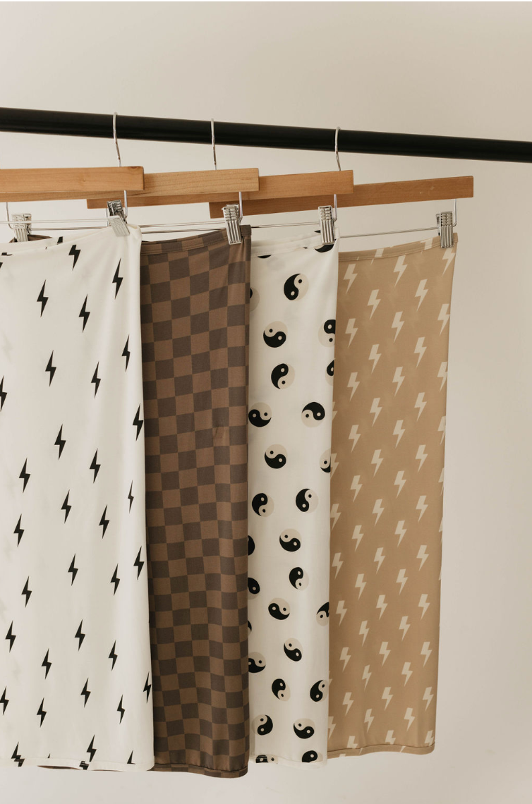 Four patterned fabrics are hanging on wooden hangers from a rod. The patterns from left to right are: black lightning bolts on white, brown checkered squares, black yin-yang symbols on white, and the "Bamboo Swaddle" with cream lightning bolts on tan by forever french baby, ideal as hypo-allergenic baby swaddles.