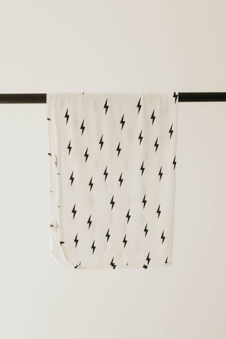 The Bamboo Swaddle in White & Black Lightning Bolt by forever french baby, featuring a white fabric with a pattern of small black lightning bolts reminiscent of breathable baby sleepwear, is draped over a black horizontal rod against a plain background.