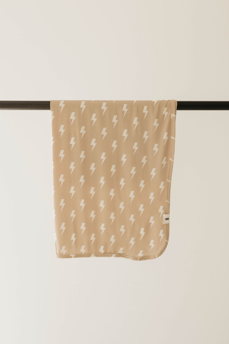 A Bamboo Swaddle from forever french baby, named Tan & Cream Lightning Bolt, embellished with a pattern of small white lightning bolts, is draped neatly over a black rod against a light background.