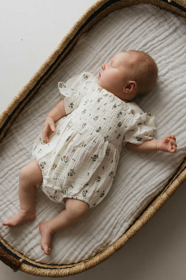 A baby sleeps peacefully in a wicker crib, dressed in the Forever French Baby Garden Days Ivory Floral Romper with short sleeves, one hand resting at their side. The crib is lined with a soft, quilted pad from the Garden Days collection by Forever French Baby. The scene is calm and serene.