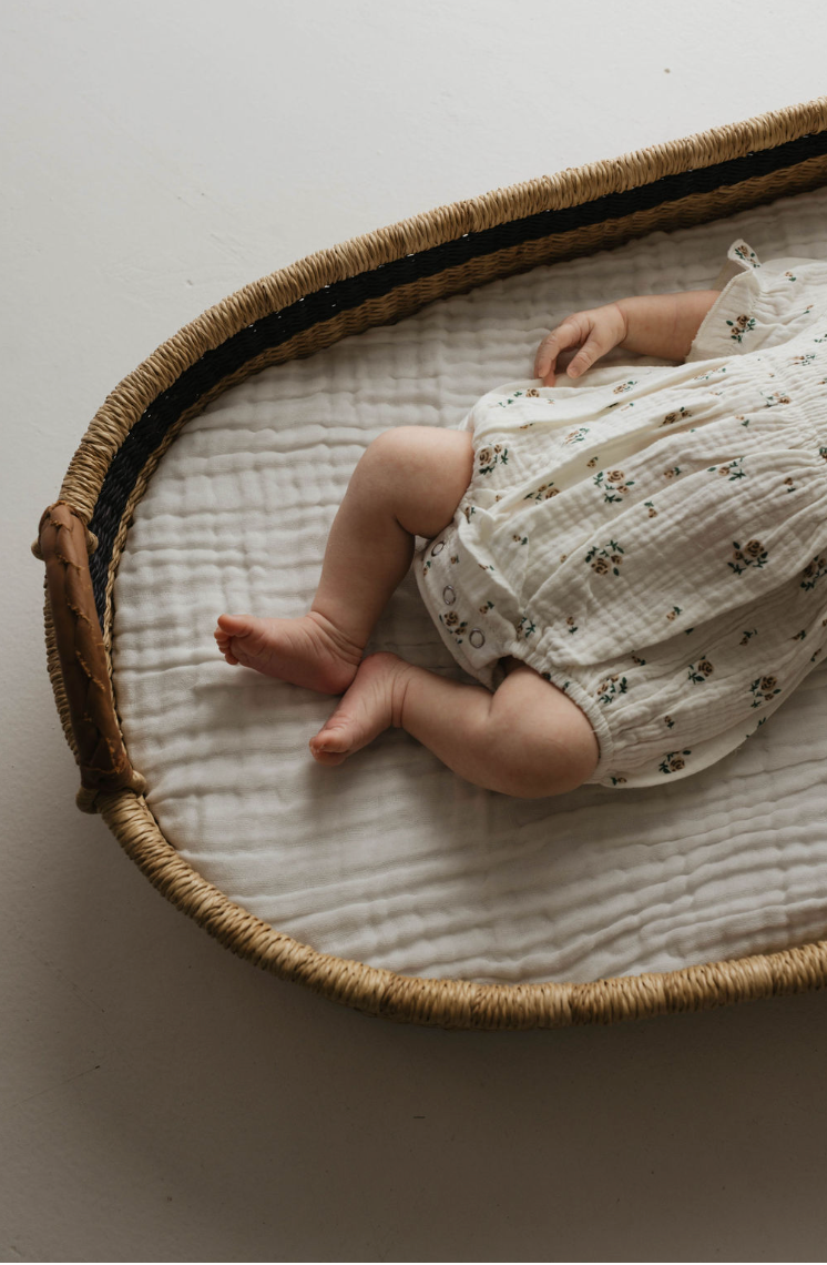 A baby lies in a woven basket, partially visible with only the legs and lower torso showing. The baby is wearing an Ivory Floral Romper from the Garden Days collection made of soft muslin fabric, resting on a white quilted mattress. The edge of the basket features a tan and black trim, adding to the cozy scene brought to you by forever french baby.