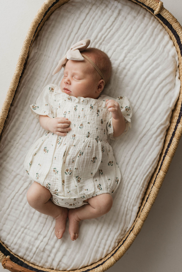 A baby sleeps in a wicker bassinet on a white blanket, snug in the Garden Days | Ivory Floral Romper from forever french baby. The bassinet has natural, woven sides and a textured mattress, perfectly complementing the baby's light-colored headband with a bow.