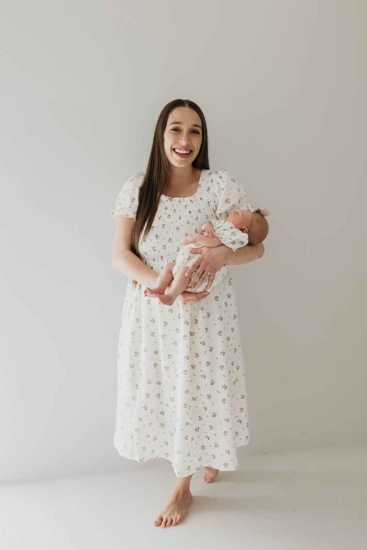 A woman with long, brown hair stands barefoot against a plain, light background. She is wearing a white dress with a floral pattern and is smiling while cradling her baby dressed in a Forever French Baby "Garden Days | Ivory Floral Romper." Both appear content and at ease.