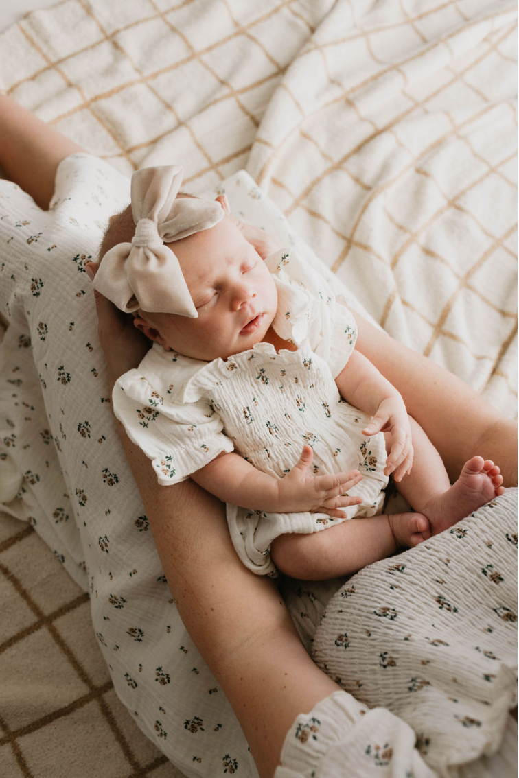A baby wearing the Garden Days | Ivory Floral Romper from forever french baby and a large beige bow headband sleeps peacefully in an adult's lap. The adult, dressed in a matching floral outfit, cradles the baby gently against a plaid blanket background.