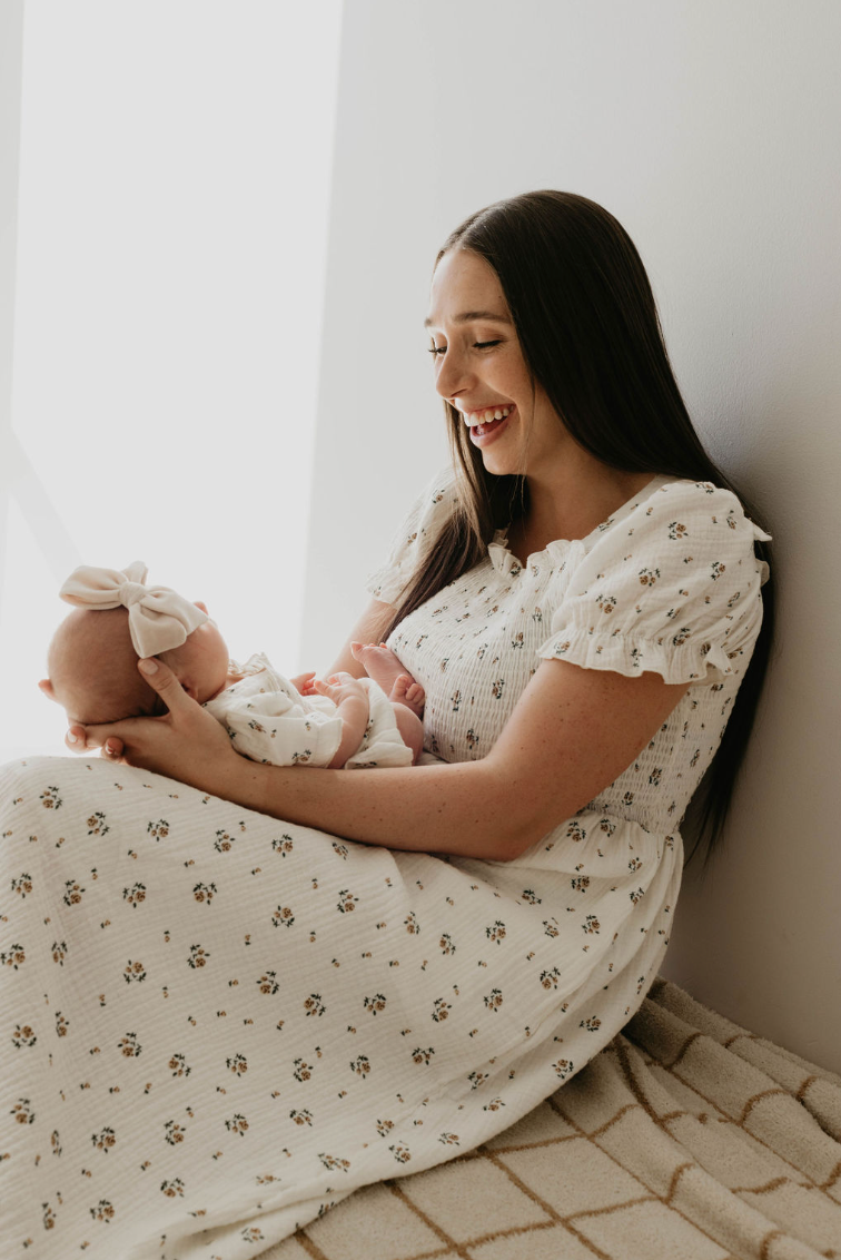 A woman with long dark hair, wearing a white floral dress, sits on the floor by a wall, holding and gazing lovingly at a baby dressed in Forever French Baby's Garden Days Ivory Floral Romper with a large bow headband. Both are bathed in soft natural light.