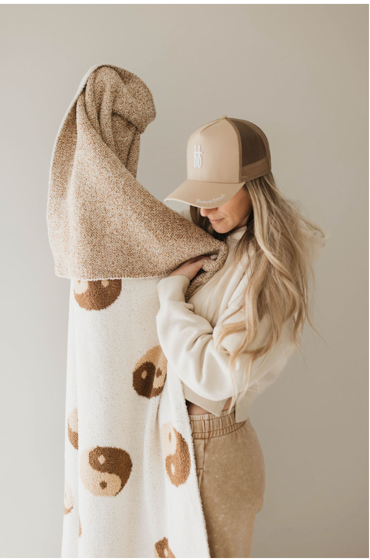 A woman with long hair, wearing a beige and brown Adult Trucker Hat by Forever French Baby and matching casual clothing, stands holding a Forever French Baby beige and white blanket with a bear pattern. The neutral background puts the focus on her and the blanket, which should be spot cleaned or washed with mild detergent.