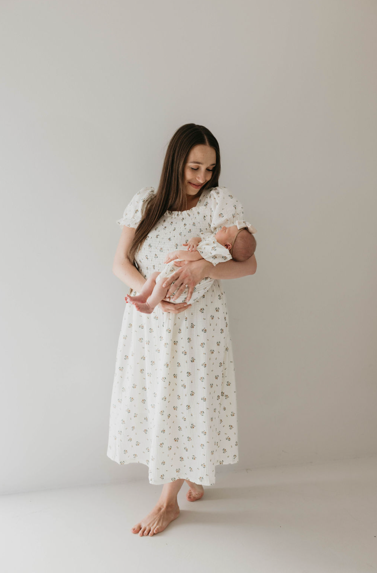 A woman stands barefoot against a plain background, gazing down as she cradles a baby. She is wearing the "Child & Women's Muslin Dress | Garden Days" by forever french baby, adorned with an ivory muslin fabric and a charming floral pattern. The baby is dressed in a matching outfit, and both appear serene and content.