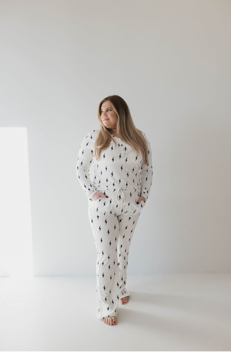 A woman stands in a minimalist white room wearing the Women's Bamboo Pajamas by forever french baby. The matching set, featuring white pajama-style pants and a long-sleeved top adorned with small black and white lightning bolt prints, exudes comfort and style as she looks slightly to her left with her hands in her pockets.