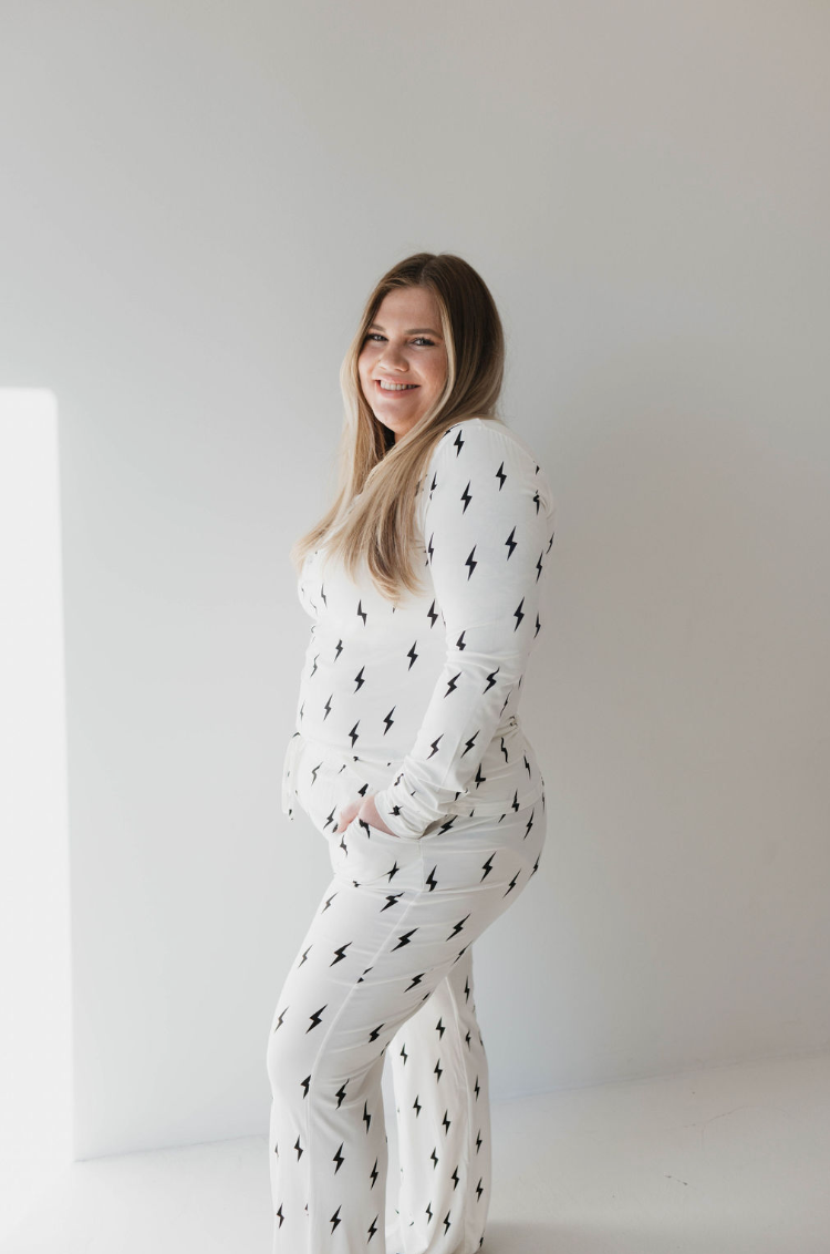 A smiling woman with long, light brown hair stands facing slightly to the left. She is wearing a set of Women's Bamboo Pajamas by forever french baby, featuring a white design with black and white lightning bolt prints. The outfit includes a long-sleeve top and pants, and her left hand is casually placed in her pocket. The background is plain white.
