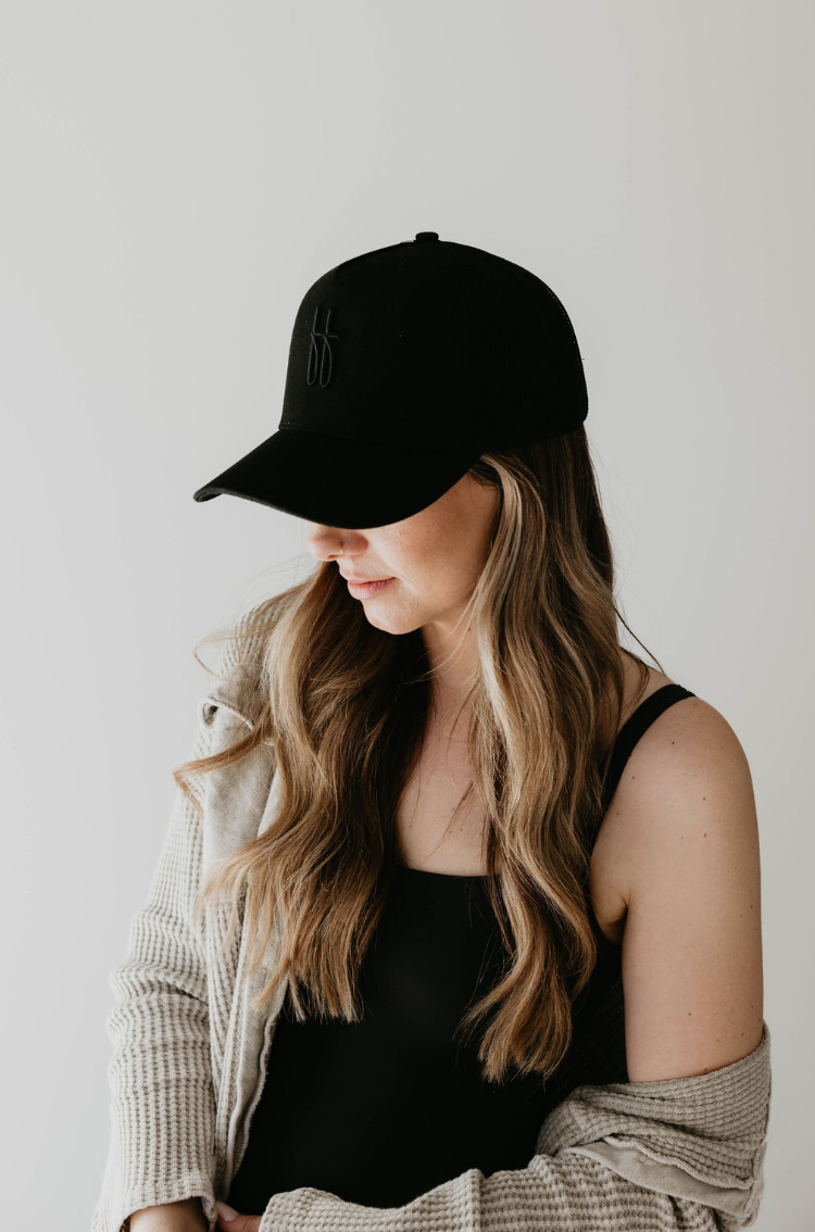 A woman with long, wavy hair wears a black tank top and the ff Tan Adult Trucker Hat by forever french baby. She has a loose-knit cardigan draped over her shoulders that can be spot cleaned with mild detergent. She is looking downwards, and the background is plain white.