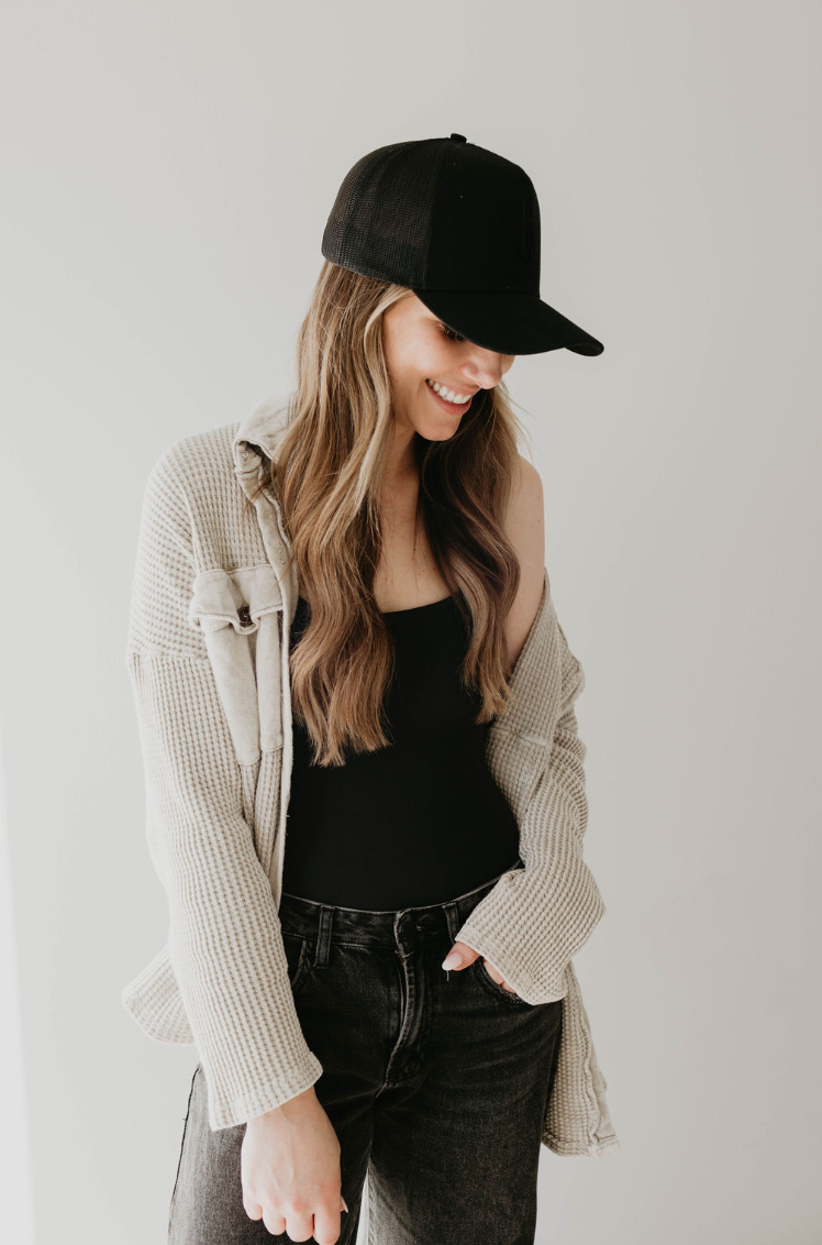 A person with long hair stands against a plain white background, facing slightly to the right. They are wearing an Adult Trucker Hat in tan from forever french baby, a beige checkered overshirt from Forever French Baby, a black tank top, and dark jeans. They are looking down and smiling.