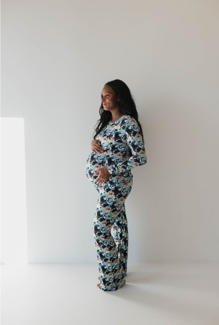 A pregnant woman stands sideways against a plain white background, gently cradling her belly with both hands. She wears the colorful camouflage-patterned Women's Bamboo Pajamas in the Charli Print from forever french baby, and she has long, dark hair flowing loosely over her shoulders.