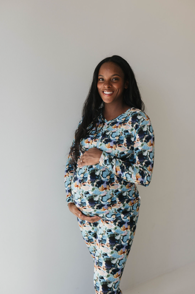 A pregnant woman with long dark hair, wearing the Charli Print bamboo pajamas from forever french baby, smiles at the camera. She holds her baby bump with both hands and stands against a plain light-colored wall in her stylish ensemble.