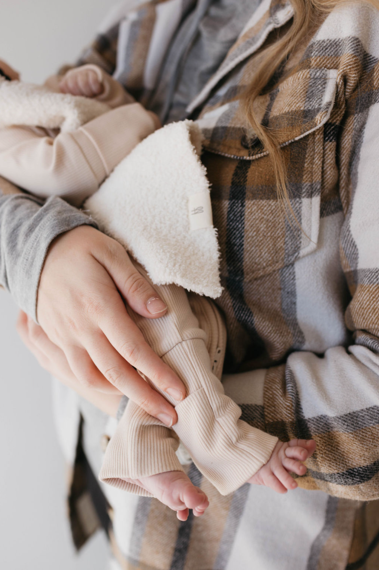 A person in a plaid jacket gently cradles a baby wearing forever french baby's Bamboo Zip Pajamas in Ribbed Cream, enhancing the loving scene with the pajamas' warm and tender touch against the baby's skin.