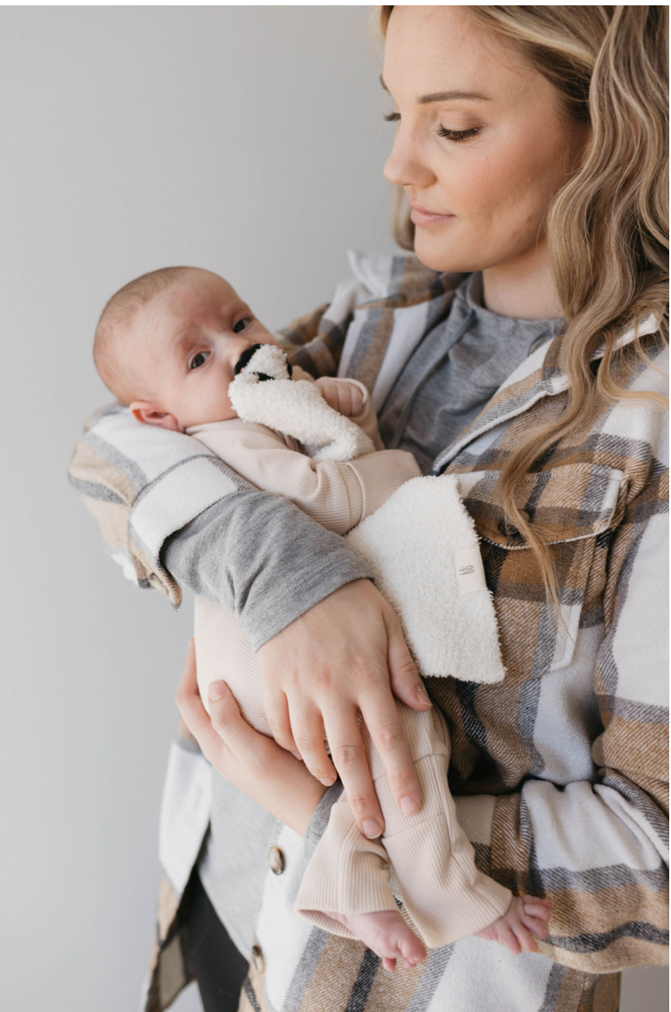 A woman with long hair holds a baby wrapped in a soft beige blanket. The baby clutches a plush toy while wearing forever french baby's Bamboo Zip Pajamas in Ribbed Cream. The woman, dressed in a plaid jacket, gazes down lovingly.