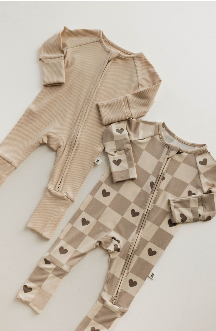 Two baby onesies from forever french baby are laid flat: Bamboo Zip Pajamas in ribbed cream with a front zipper and another in beige and brown checkered pattern with heart motifs. Both made of soft bamboo fabric, featuring long sleeves and footed legs for cozy comfort.