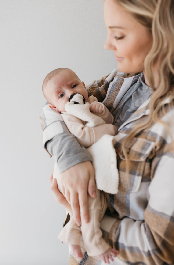 A woman in a plaid shirt gently holds her baby, wrapped in a forever french baby ribbed cream blanket, wearing adorable Bamboo Zip Pajamas. The baby clutches a plush toy while she smiles softly against a plain white wall.