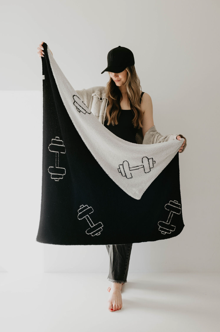 A person wearing a black hat and black tank top stands barefoot on a white surface, holding the "Plush Blanket | Arm Day" from Forever French Baby, which features dumbbell designs. They have a light-colored cardigan draped over their shoulders from Forever French Baby and are smiling while looking at the blanket.