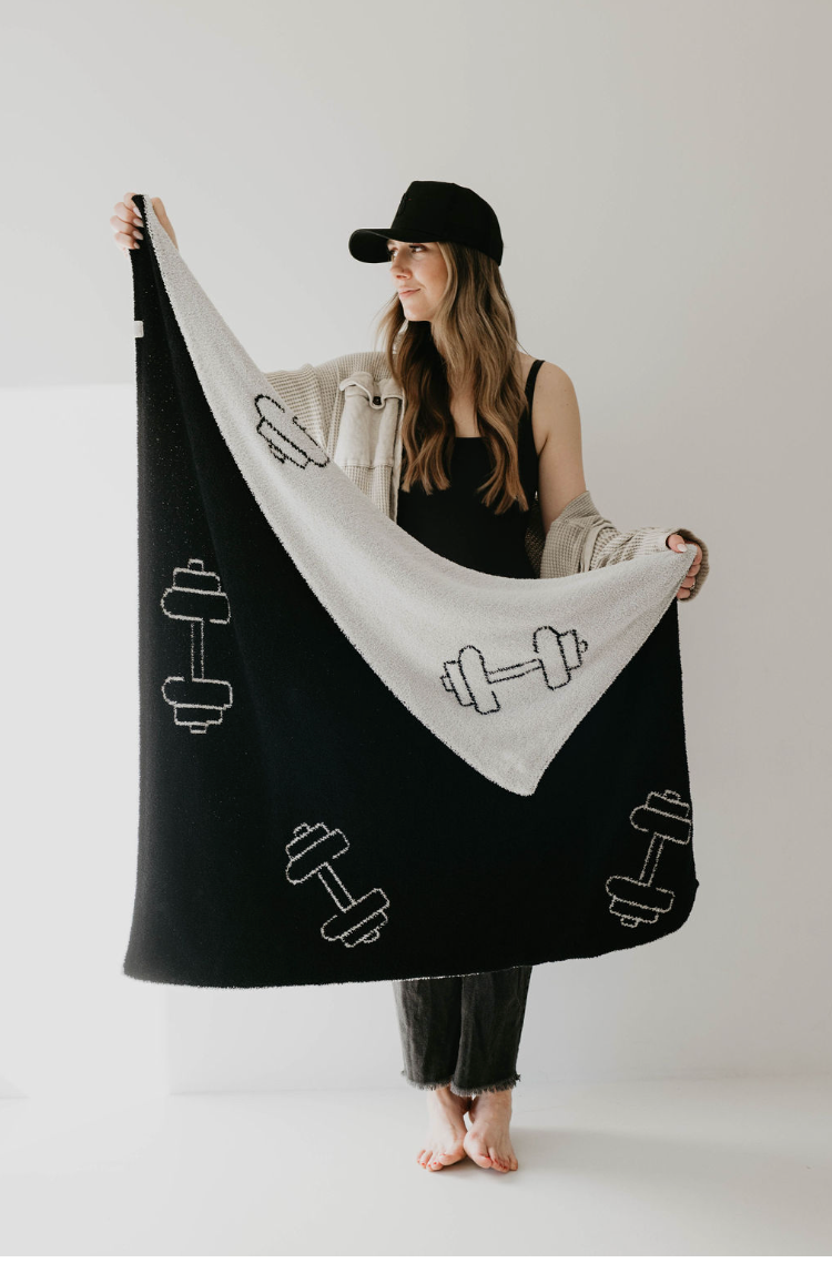 A woman in a black cap, black tank top, and light-colored cardigan holds up the "Plush Blanket | Arm Day" by forever french baby. The Oeko-tex certified blanket, made from microfiber feather yarn, showcases a pattern of dumbbells in black and white. She stands barefoot on a white floor against a plain white background.