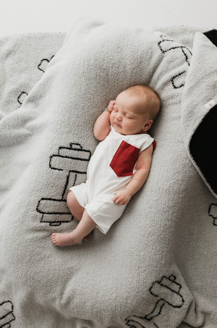 A baby dressed in a white Forever French Baby outfit with a red pocket sleeps on a soft Plush Blanket | Arm Day by forever french baby, which is Oeko-tex certified and features black and white barbell designs. The baby is lying on their back with one hand near their face and the other hand resting on the blanket.