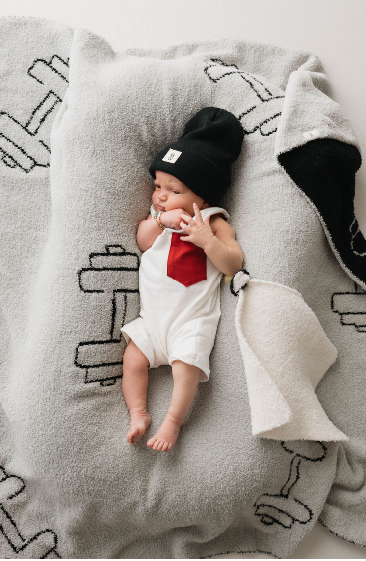 A baby lies on a "Plush Blanket | Arm Day" from forever french baby, which is Oeko-tex certified and features black and white bone patterns on a gray background. The baby is dressed in a white onesie with a red tie design and is wearing a black beanie. One corner of the soft microfiber feather yarn blanket is folded over, partially covering the baby's legs.