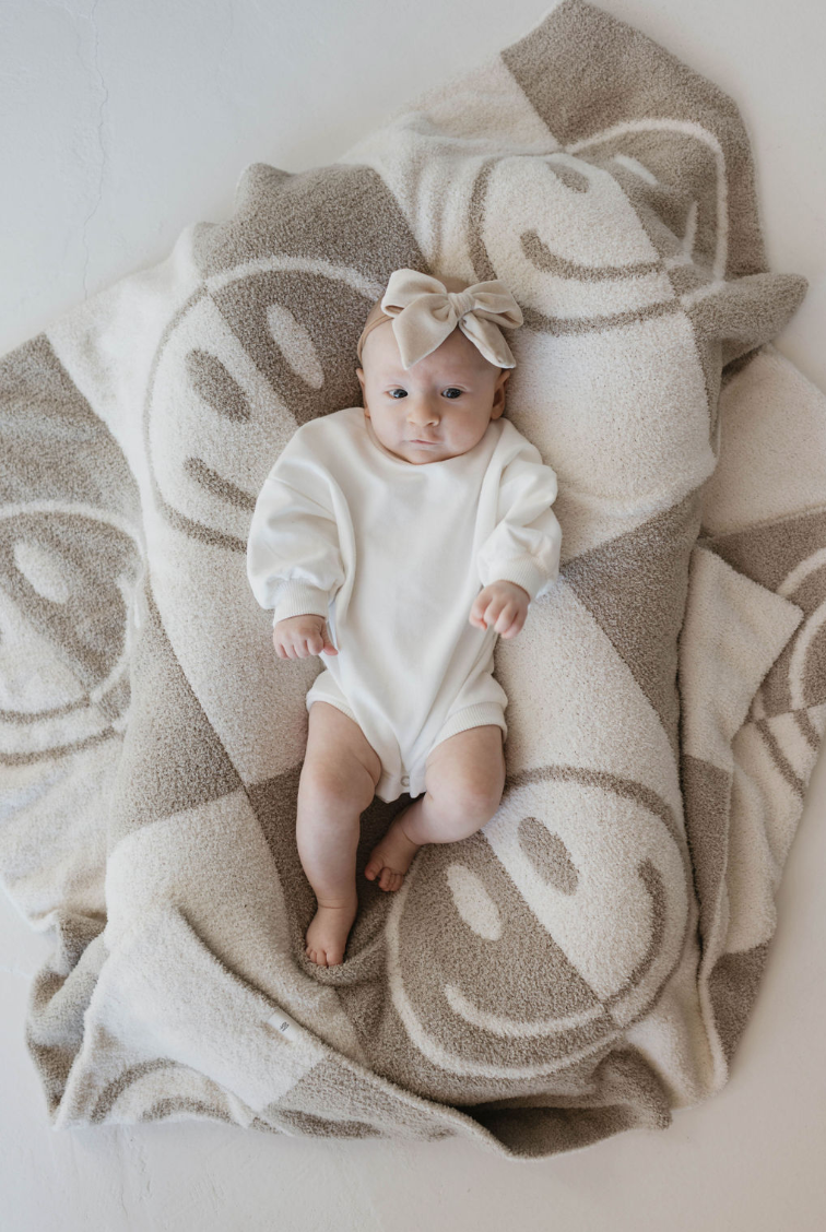 A baby wearing a white onesie and a beige bow headband lies on a fuzzy, beige and white OEKO Standard 100 Plush Blanket | Smile Checkerboard by forever french baby. The baby looks up with wide eyes, her arms resting by her sides. The blanket is spread out, creating a cozy and soft environment.