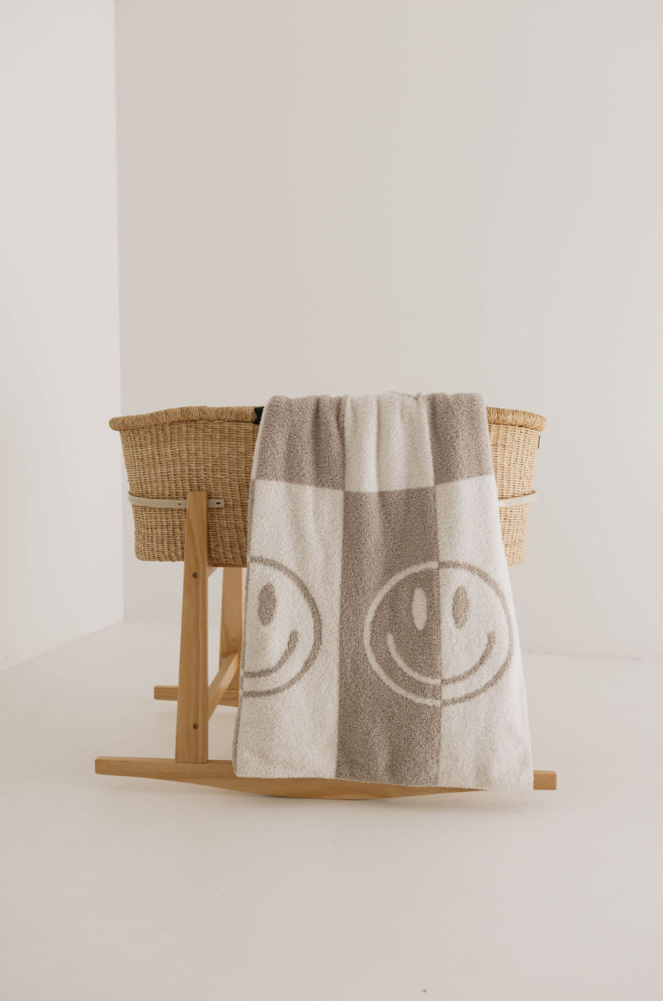 A Plush Blanket | Smile Checkerboard by forever french baby hangs over the side of a wooden, basket-like stand against a plain white background, reminiscent of a cozy baby blanket, displaying two large smiling faces.