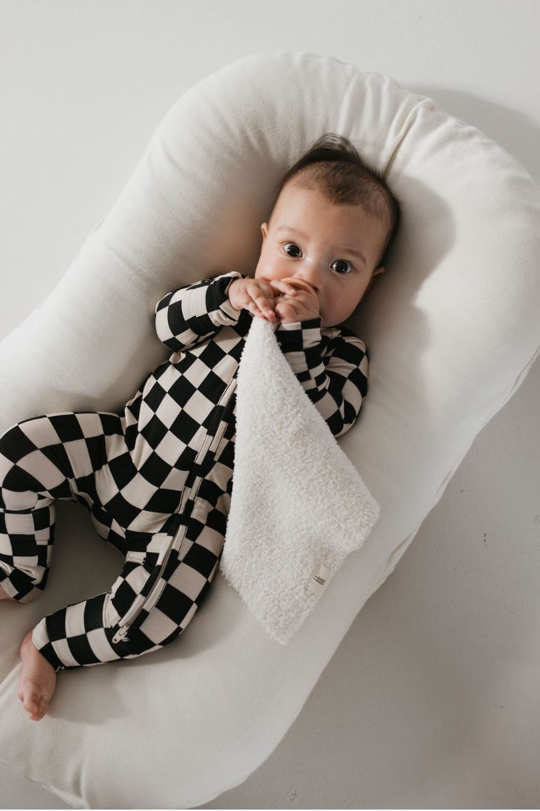 A baby wearing Bamboo Zip Pajamas in a Black Checkerboard pattern from forever french baby is lying on a white pillow. The baby is holding a white textured cloth close to their face, looking up with wide eyes. The background is a plain, light-colored surface.