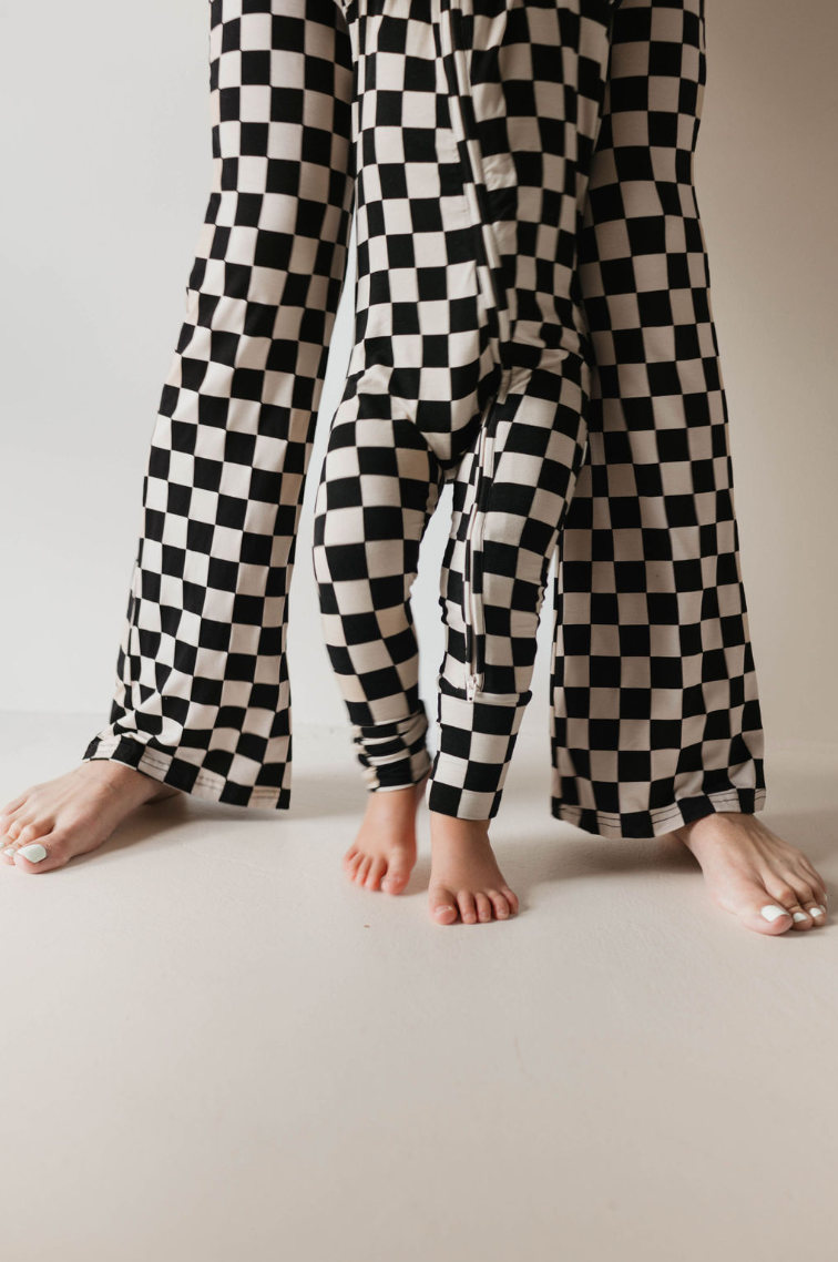A woman and a child, both dressed in matching "Women's Bamboo Pajamas | Black Checkerboard" from forever french baby, are standing together. The woman's feet are pointed forward while the child's feet are slightly turned. The focus is on their pajama pants and bare feet against a neutral background.